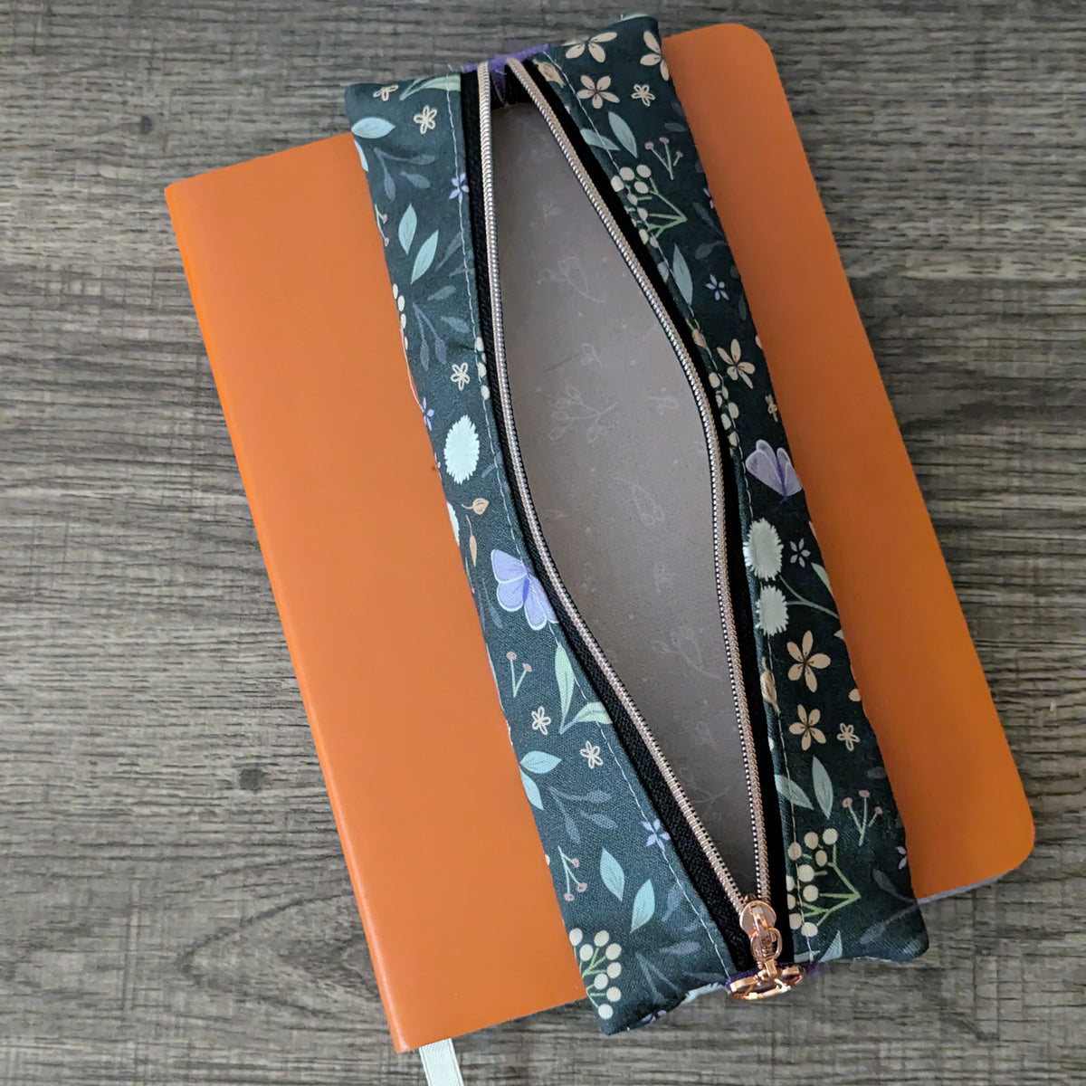 A dark teal floral zipper pouch with a rose gold zipper, shown open and laid diagonally over an orange notebook on a gray wood-textured background.