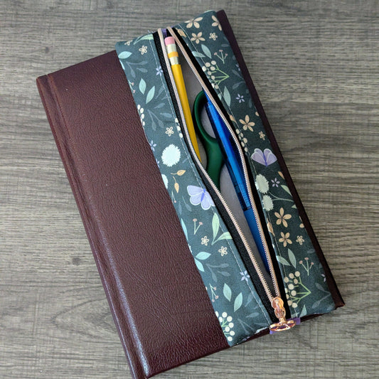 A floral zippered pouch with a gold zipper, shown open next to a brown notebook on a gray wood-grain surface. The pouch features a green background with white and purple flowers, and contains a yellow pencil, green scissors, and a blue pen.