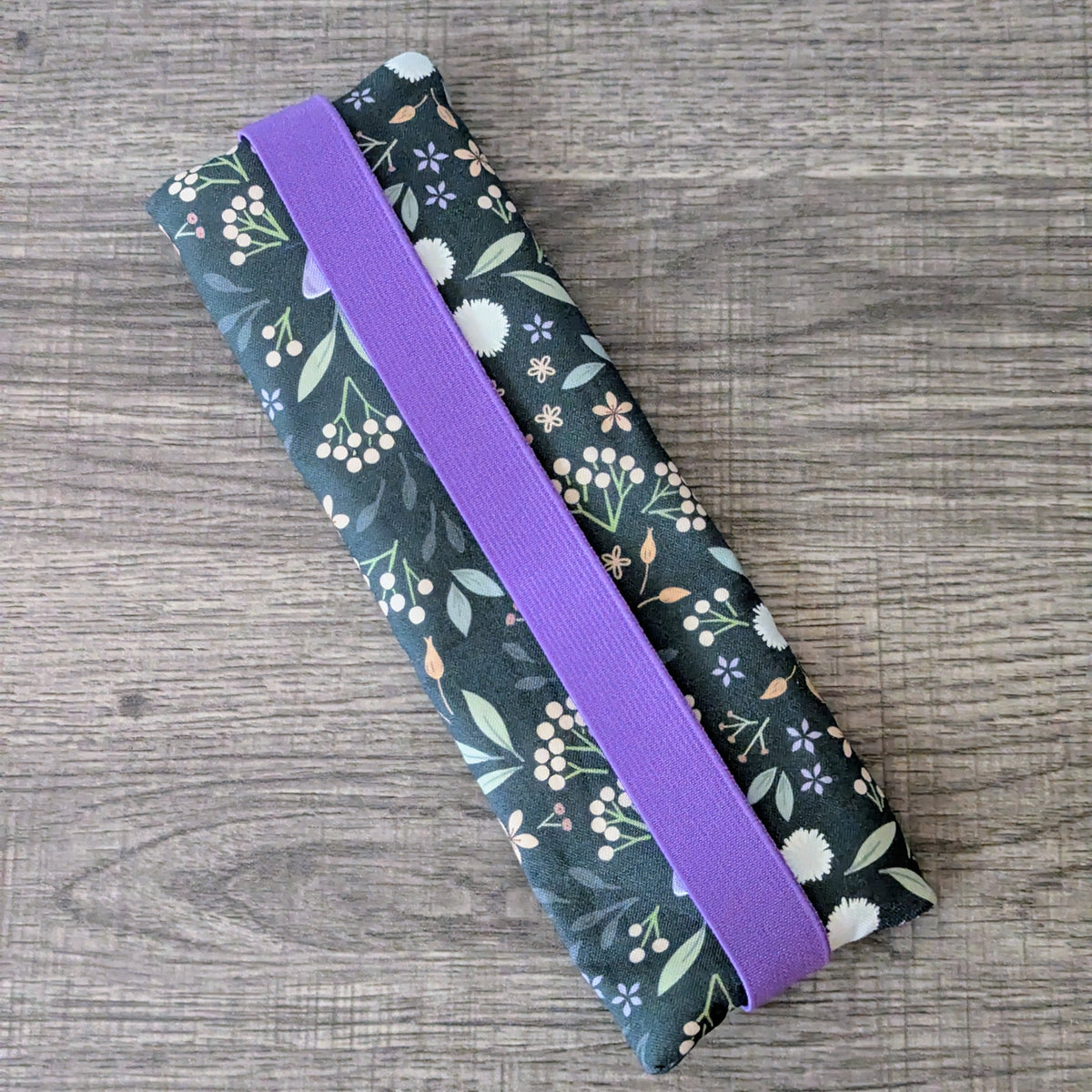 A rectangular fabric pouch with a dark green floral print and a wide purple elastic band, photographed on a gray wood-textured surface.