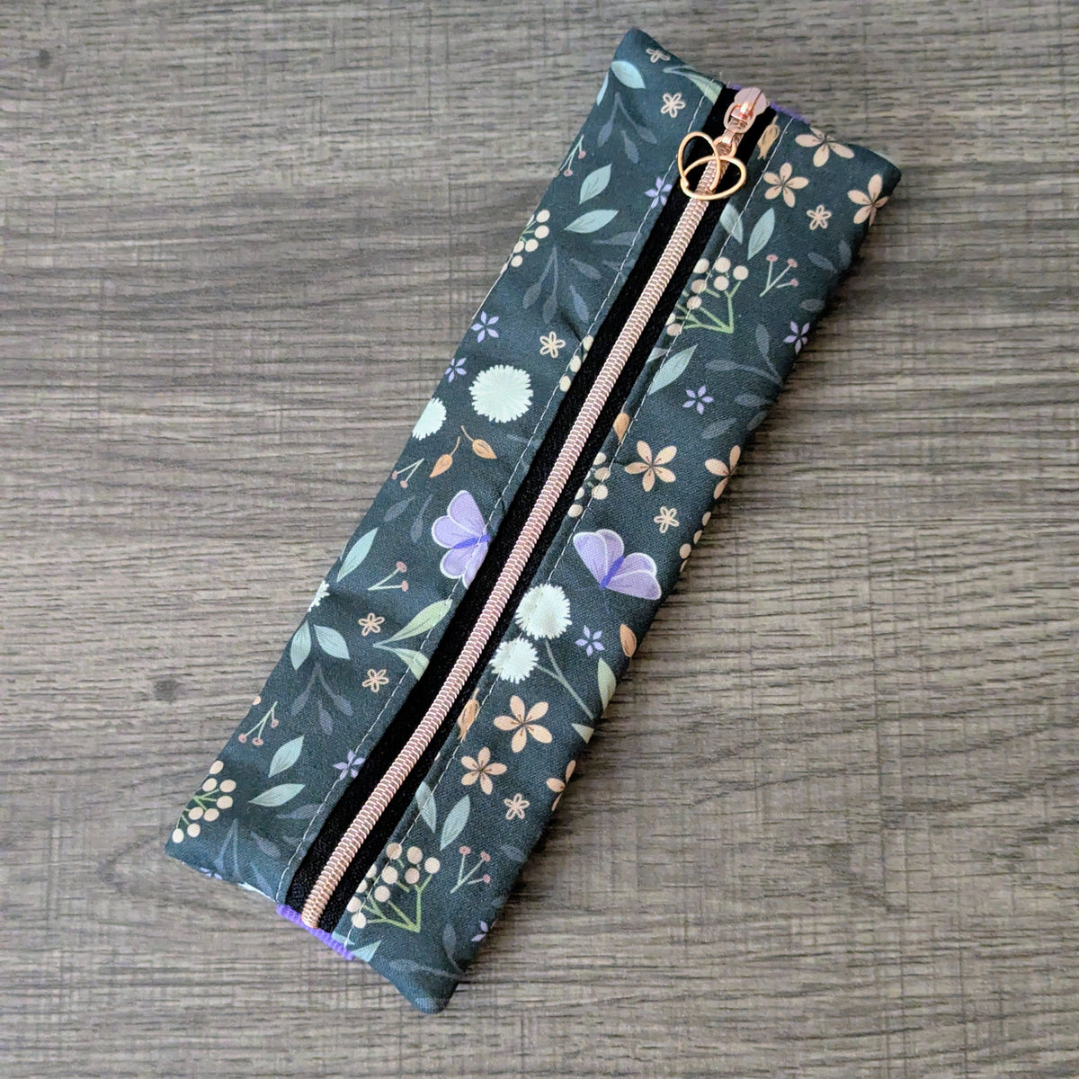 A rectangular zippered pouch with a dark teal floral print, featuring purple butterflies, white flowers, and a rose gold zipper pull, shown on a gray wood-textured background.