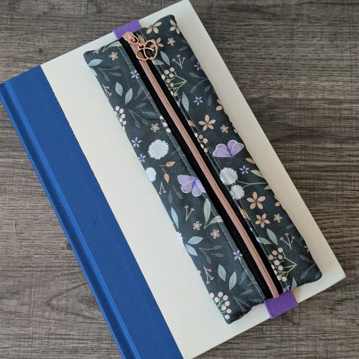 A floral zippered pencil case with a gold-tone zipper and purple elastic bands, resting diagonally on top of a blue and white notebook over a gray wood-grain surface.