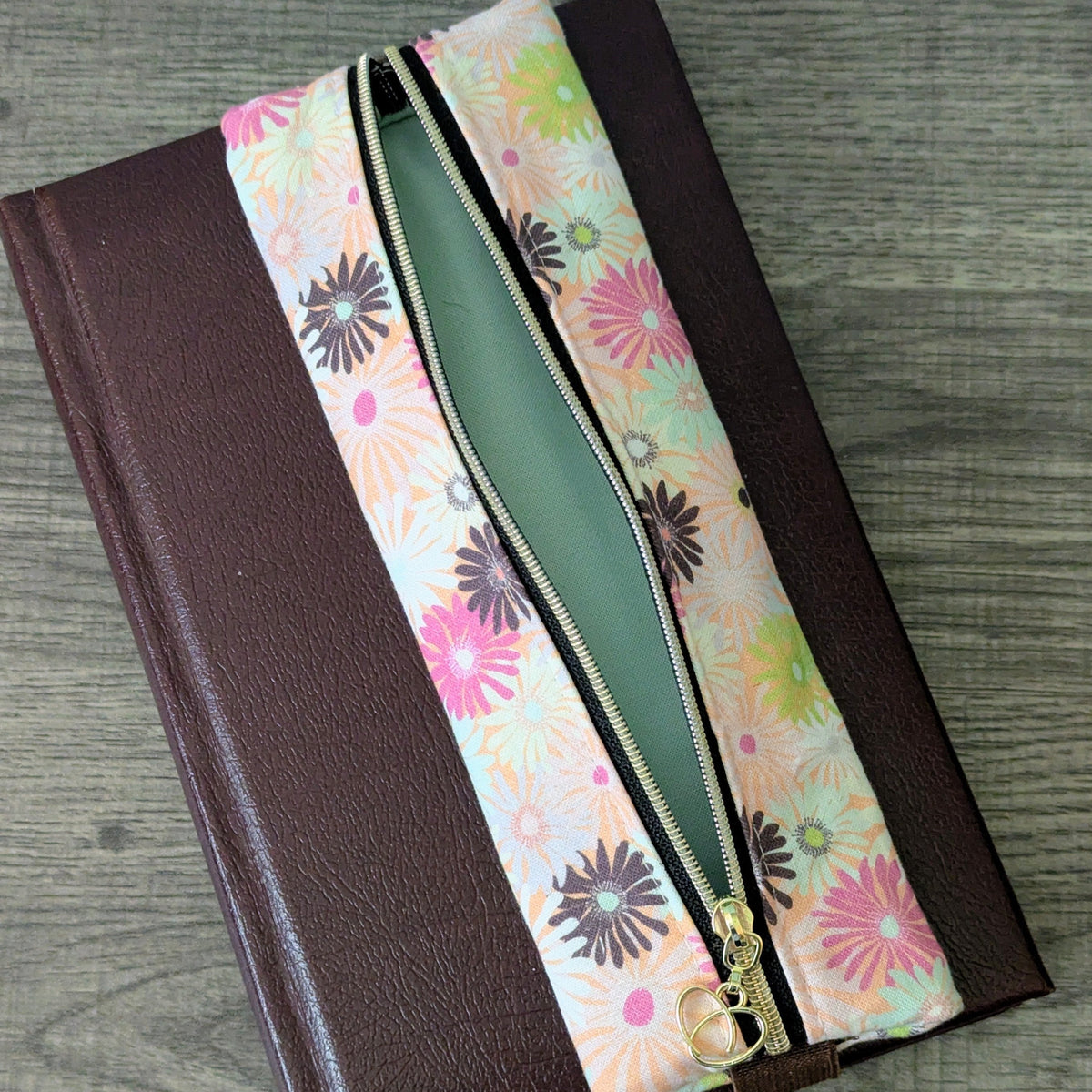 A brown faux-leather journal with a zippered pouch with a colorful floral patterned fabric, shown open on a gray wood-textured surface.