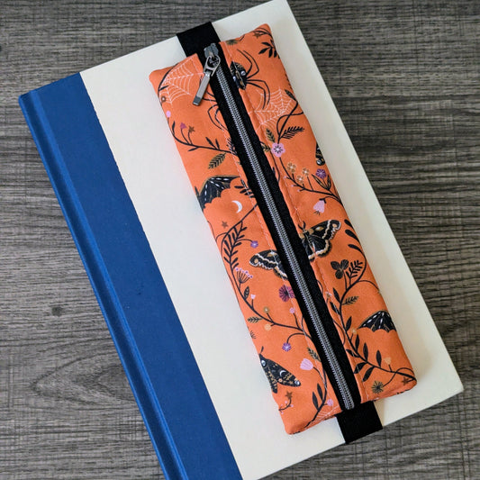 An orange zippered pencil case with a black and white butterfly and floral pattern, resting on a white notebook with a blue spine on a gray wood-textured surface.