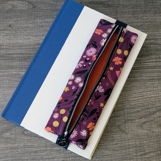 A floral zippered pouch with a purple base and multicolor flower print, laid diagonally across a blue and white striped notebook on a gray wood-grain surface.