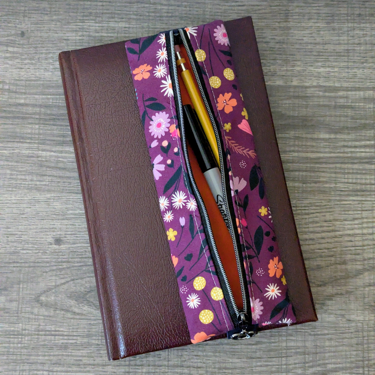 A brown hardcover notebook with a floral-patterned zipper cover, shown open on a gray wood-grain surface. Inside the cover, yellow and black pens are visible.