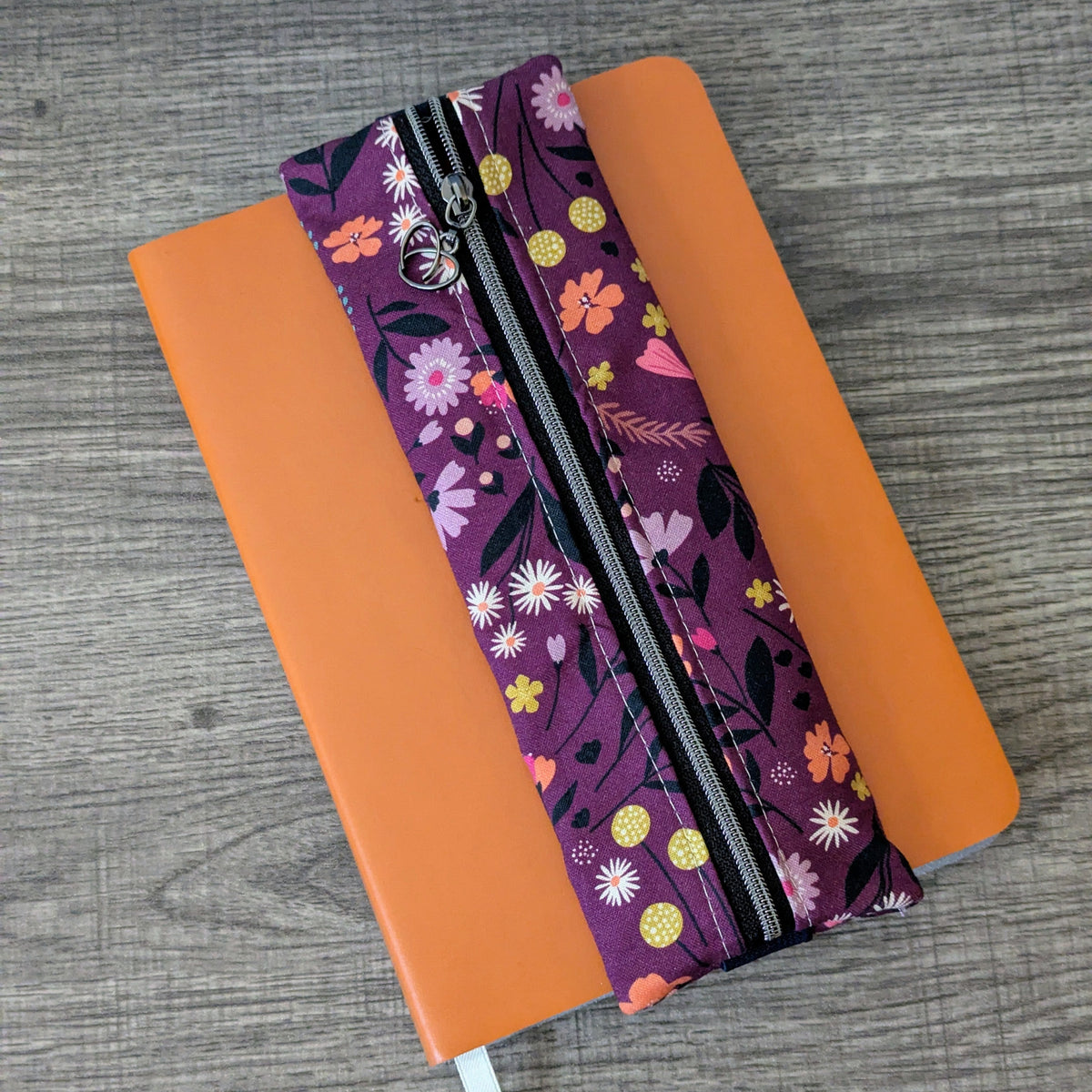 An orange hardcover notebook with a white ribbon bookmark, featuring a purple floral zipper pouch attached along the spine, placed on a gray wood-textured surface.