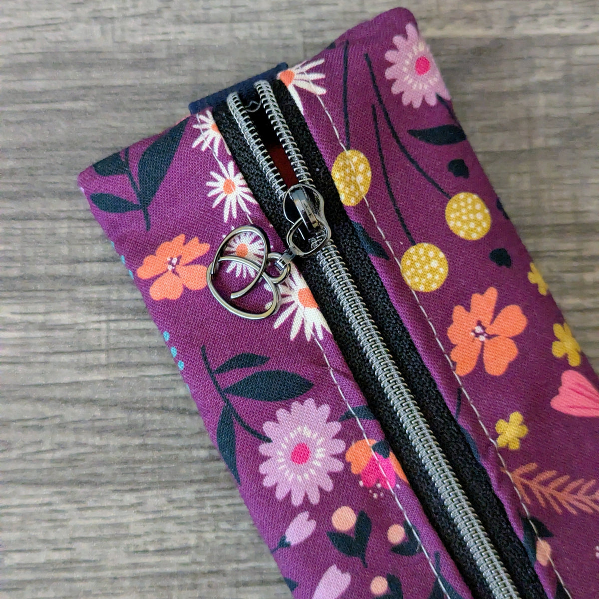 A close-up of a purple fabric zipper pouch with a colorful floral print, featuring orange, pink, and yellow flowers with black leaves. The pouch is shown partially open, revealing a black zipper with a silver metal pull tab shaped like a heart.