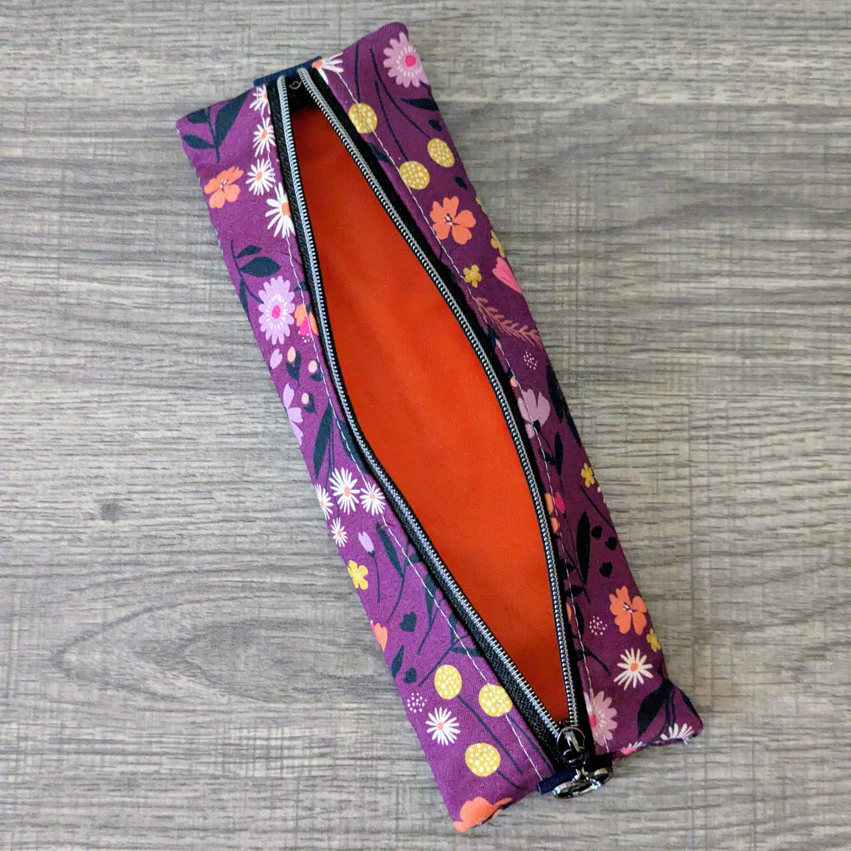 A rectangular zippered pouch with a purple floral pattern exterior and a bright orange interior, shown open on a gray wood-textured background.