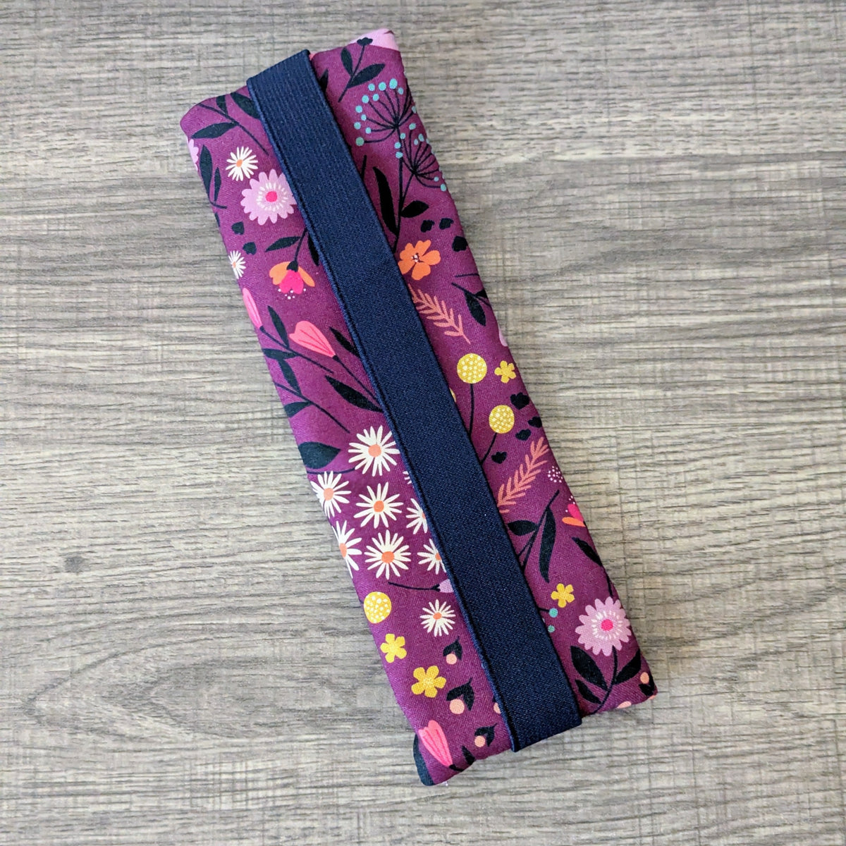 A fabric pouch with a purple floral print and a navy blue elastic band, shown on a gray wood-textured background.
