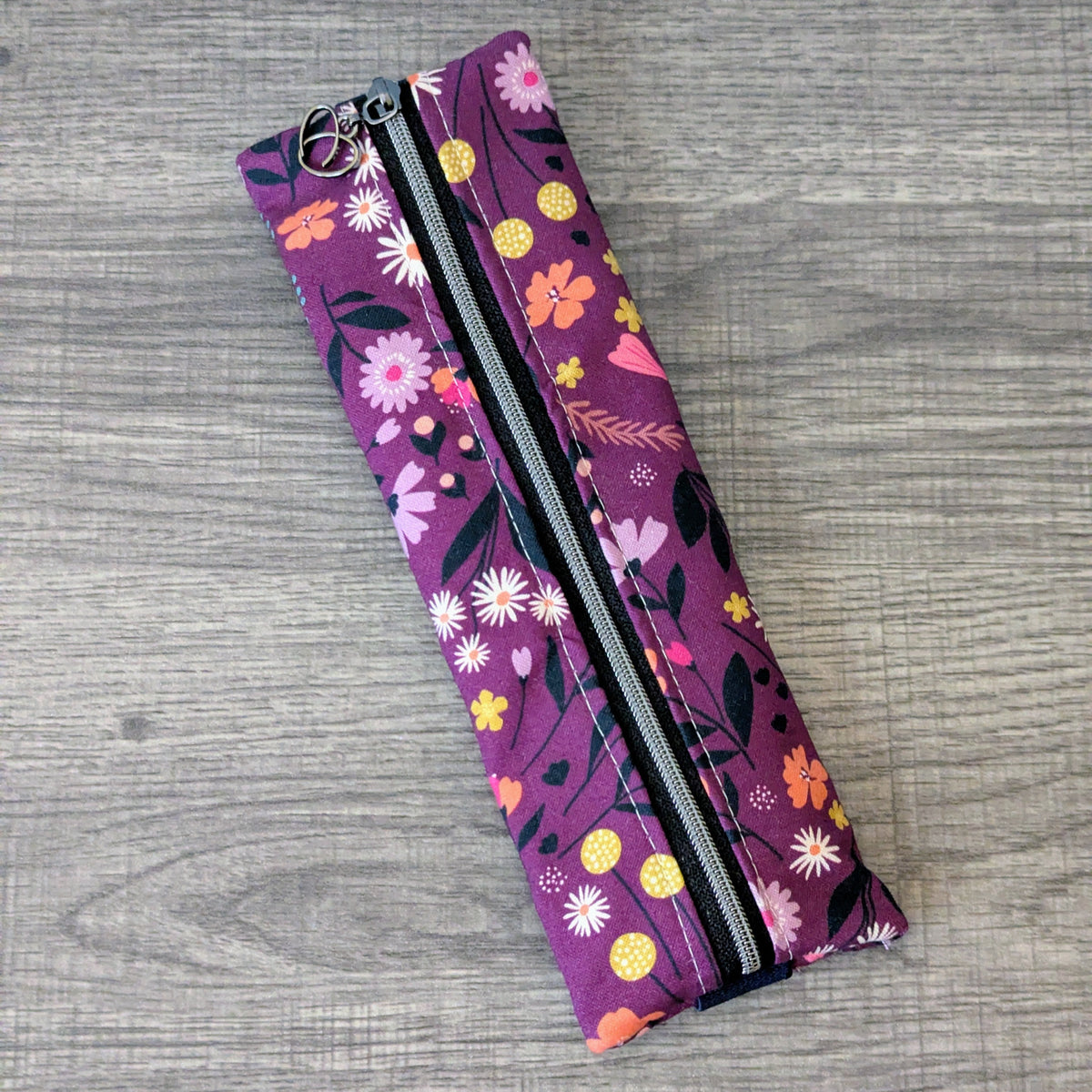 A rectangular zippered pouch with a purple floral print, featuring pink, yellow, and white flowers with green leaves, laid flat on a gray wood-textured surface.