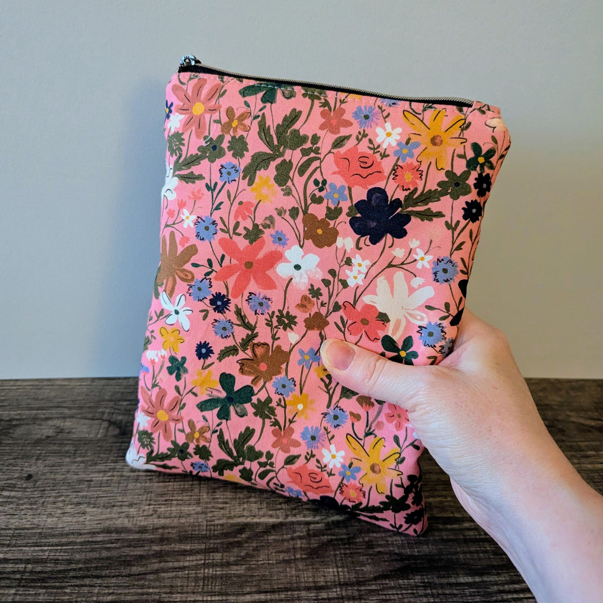 Floral-patterned pouch held by a hand on a wooden surface with a neutral background