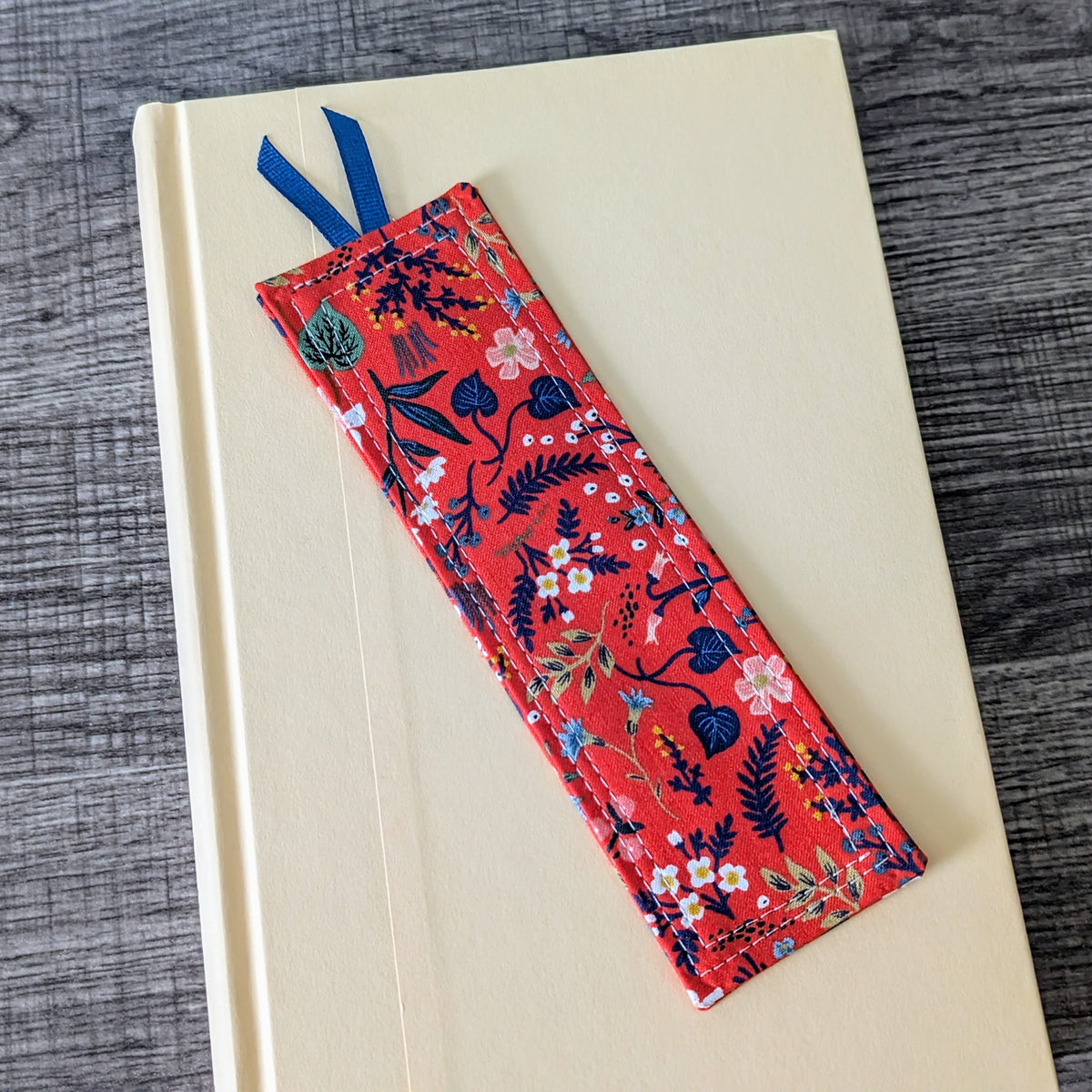 A red cotton bookmark with a wild flower print, featuring a coordinating ribbon, placed on an closed book.